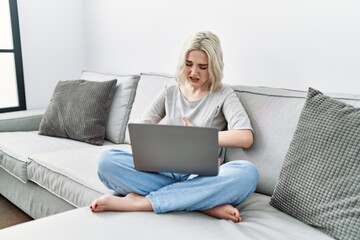 Young caucasian woman using laptop at home sitting on the sofa with hand on stomach because indigestion, painful illness feeling unwell. ache concept.