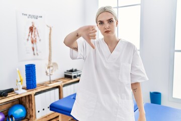 Young caucasian woman working at pain recovery clinic looking unhappy and angry showing rejection and negative with thumbs down gesture. bad expression.