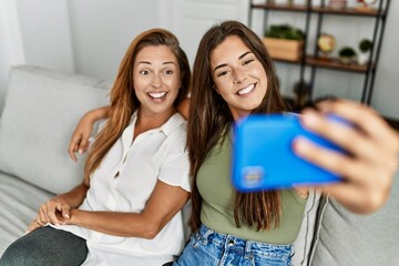 Mother and daughter smiling confident and hugging each other making selfie by the smartphone at home