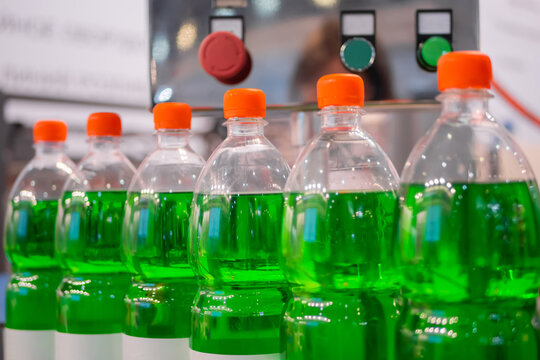 Row Of Pet Bottles With Green Lemonade And Orange Caps On Conveyor Belt Of Automatic Liquid Filling Machine At Plastic Exhibition - Close Up. Manufacturing, Industry And Technology Equipment Concept