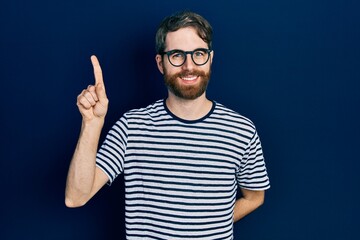 Fototapeta premium Caucasian man with beard wearing striped t shirt and glasses showing and pointing up with finger number one while smiling confident and happy.