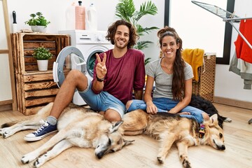 Young hispanic couple doing laundry with dogs showing and pointing up with fingers number two while smiling confident and happy.