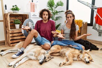 Young hispanic couple doing laundry with dogs clueless and confused with open arms, no idea concept.