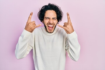 Handsome hispanic man wearing casual white sweater shouting with crazy expression doing rock symbol...