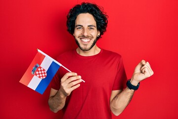 Handsome hispanic man holding croatia flag screaming proud, celebrating victory and success very...