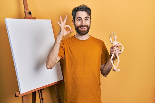 Caucasian man with beard standing by painter easel stand holding manikin doing ok sign with fingers, smiling friendly gesturing excellent symbol