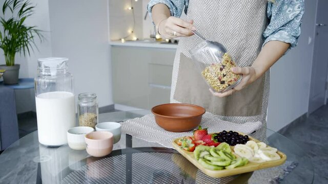 Faceless woman prepare healthy gluten free breakfast on table in kitchen. Close-up hands with spoon filling the bowl oatmeal flakes with dried fruits on large glass 