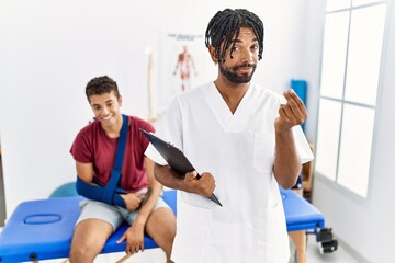 Young hispanic man working at pain recovery clinic with a man with broken arm doing money gesture...