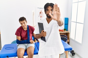Young hispanic man working at pain recovery clinic with a man with broken arm doing stop sing with palm of the hand. warning expression with negative and serious gesture on the face.