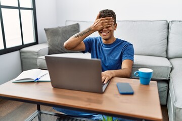Young handsome hispanic man using laptop sitting on the floor smiling and laughing with hand on face covering eyes for surprise. blind concept.