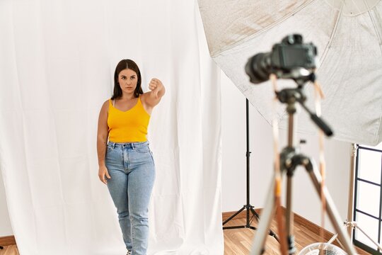 Young beautiful hispanic woman posing as model at photography studio looking unhappy and angry showing rejection and negative with thumbs down gesture. bad expression.
