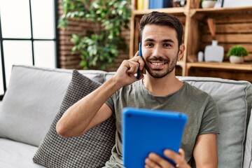 Young hispanic man using touchpad and talking on the smartphone at home