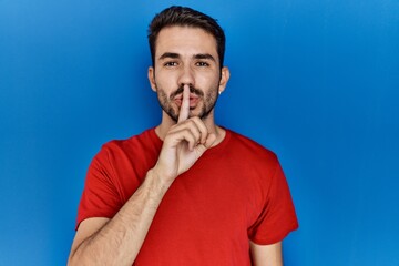 Young hispanic man with beard wearing red t shirt over blue background asking to be quiet with finger on lips. silence and secret concept.