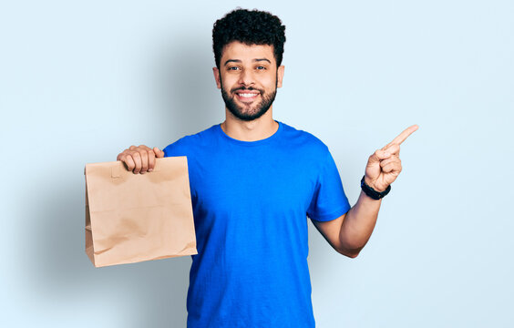 Young arab man with beard holding take away paper bag smiling happy pointing with hand and finger to the side