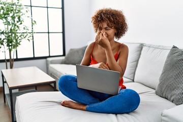 Young african american woman sitting on the sofa at home using laptop smelling something stinky and disgusting, intolerable smell, holding breath with fingers on nose. bad smell - obrazy, fototapety, plakaty