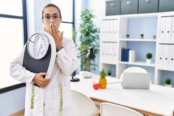 Young hispanic woman as nutritionist doctor holding weighing machine covering mouth with hand,...