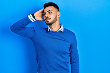 Young hispanic man with beard wearing casual blue sweater smiling confident touching hair with hand up gesture, posing attractive and fashionable