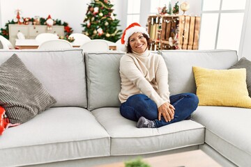 Middle age hispanic woman smiling happy wearing christmas hat sitting on the sofa at home.