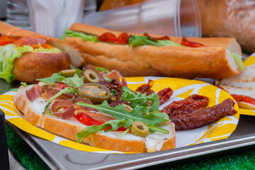 Sandwich with arugula, ham, cheese, jalapeno, sundried tomato and salami on plate on counter at summer local food market - close up. Gastronomy, street food, mediterranean and Italian dish concept
