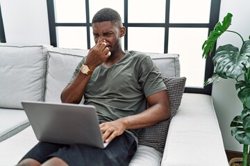 Young african american man using laptop at home sitting on the sofa smelling something stinky and disgusting, intolerable smell, holding breath with fingers on nose. bad smell - obrazy, fototapety, plakaty
