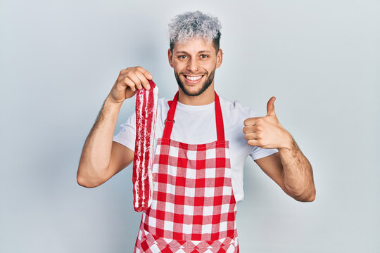 Young hispanic man with modern dyed hair wearing apron holding raw beef steak smiling happy and positive, thumb up doing excellent and approval sign