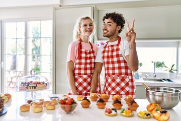 Couple of wife and husband cooking pastries at the kitchen showing and pointing up with fingers number two while smiling confident and happy.