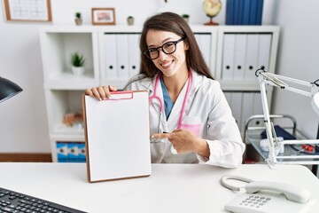 Young doctor woman holding clipboard at the clinic smiling happy pointing with hand and finger