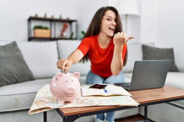 Obraz na płótnie Canvas Young brunette woman putting euro coin in piggy bank saving for travel pointing thumb up to the side smiling happy with open mouth