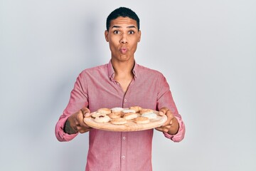 Young african american guy holding tray with cake sweets making fish face with mouth and squinting eyes, crazy and comical.