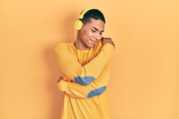 Young african american guy listening to music using headphones hugging oneself happy and positive,...