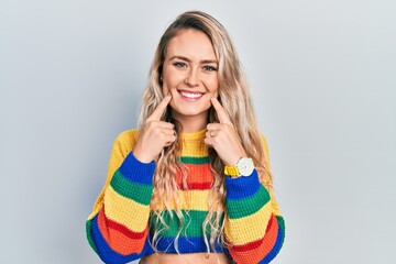 Beautiful young blonde woman wearing colored sweater smiling with open mouth, fingers pointing and...