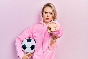 Beautiful young blonde woman holding soccer ball pointing with finger to the camera and to you, confident gesture looking serious