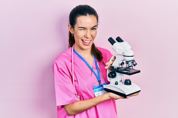 Young brunette woman holding microscope winking looking at the camera with sexy expression,...