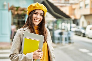 Young hispanic girl smiling happy holding laptop and book at the city.