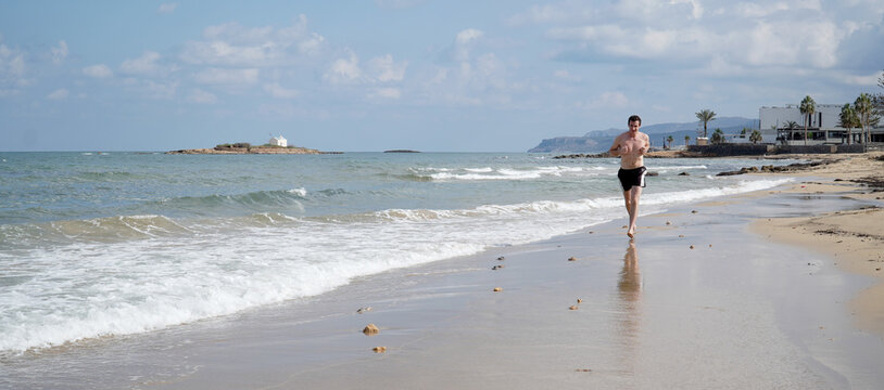 Young man jogging on a sandy beach on a sunny day in Malia, Crete Island of Greece.