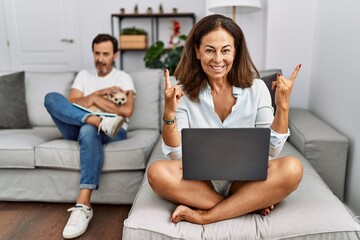 Hispanic middle age couple at home, woman using laptop shouting with crazy expression doing rock...