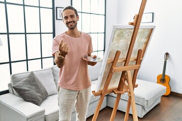Young hispanic man with beard painting on canvas at home beckoning come here gesture with hand inviting welcoming happy and smiling