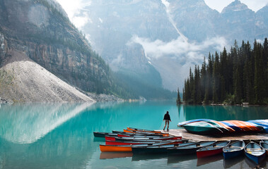 Fototapeta Tiny person standing on dock with colourful canoes at turquoise blue lake surrounded by mountains obraz