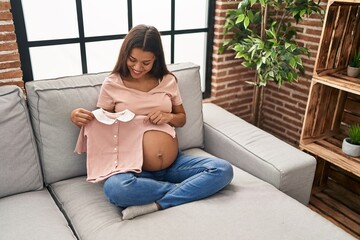 Young latin woman pregnant holding baby clothes at home