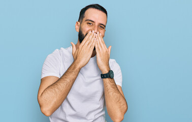 Young man with beard wearing casual white t shirt laughing and embarrassed giggle covering mouth...