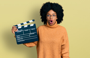 Young african american woman holding video film clapboard scared and amazed with open mouth for...