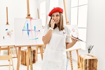 Young artist woman painting on a canvas at art studio looking stressed and nervous with hands on mouth biting nails. anxiety problem.