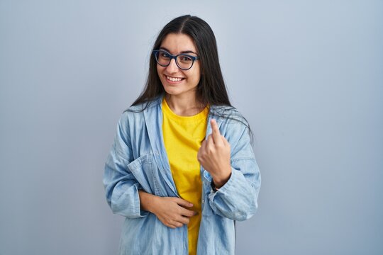 Young hispanic woman standing over blue background beckoning come here gesture with hand inviting welcoming happy and smiling