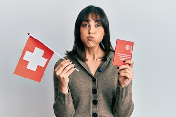 Young hispanic woman holding swiss flag and passport puffing cheeks with funny face. mouth inflated...