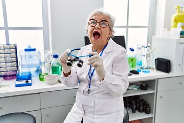 Senior woman with grey hair working at scientist laboratory using magnifying glasses angry and mad screaming frustrated and furious, shouting with anger looking up.