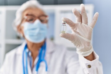 Senior grey-haired woman wearing doctor uniform and medical mask holding vaccine dose at clinic