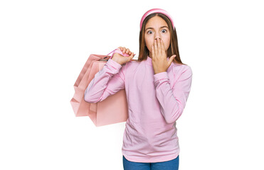 Beautiful brunette little girl holding shopping bags covering mouth with hand, shocked and afraid...