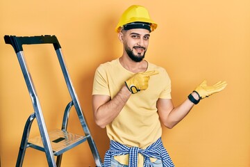 Handsome man with beard by construction stairs wearing hardhat amazed and smiling to the camera while presenting with hand and pointing with finger.