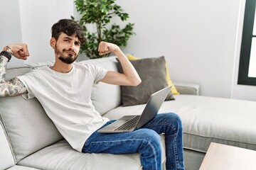 Hispanic man with beard sitting on the sofa showing arms muscles smiling proud. fitness concept.