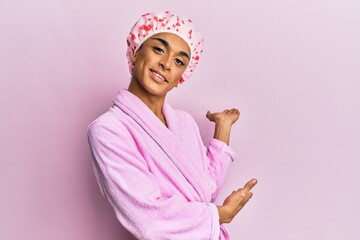 Hispanic man wearing make up wearing shower towel cap and bathrobe inviting to enter smiling...
